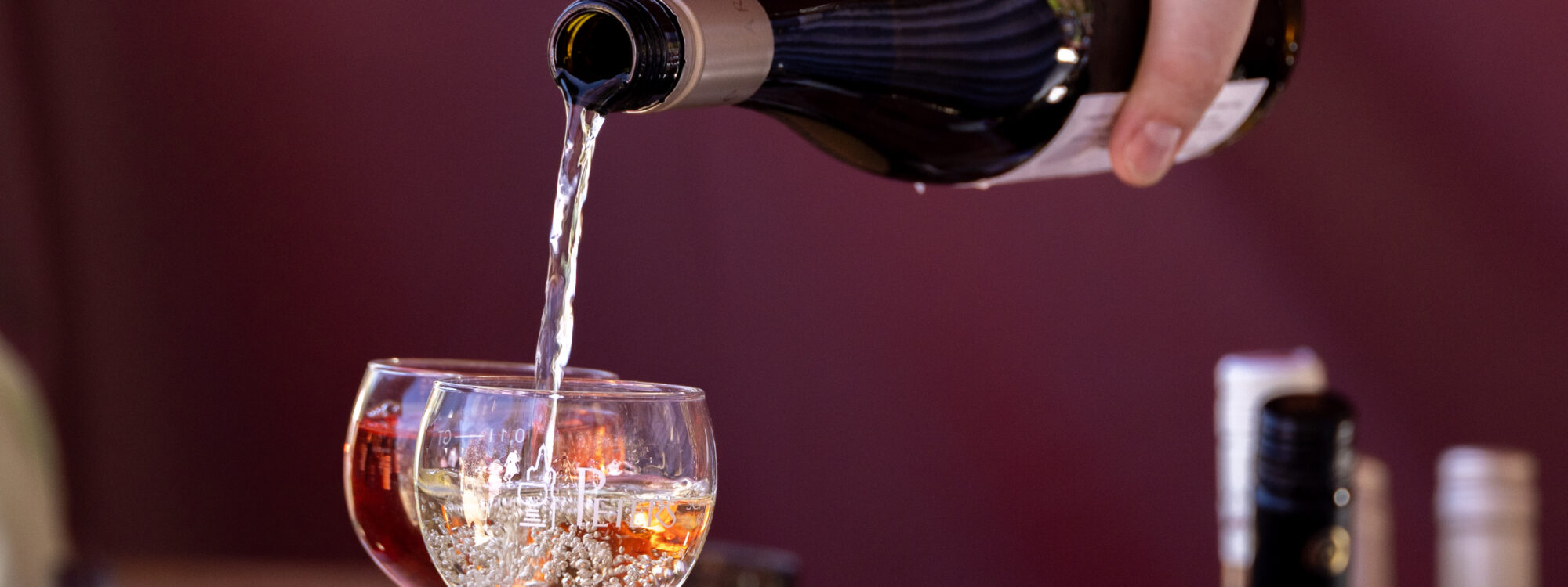 Eine Hand schüttet aus einer Glasflasche eine Flüssigkeit in zwei Gläser.