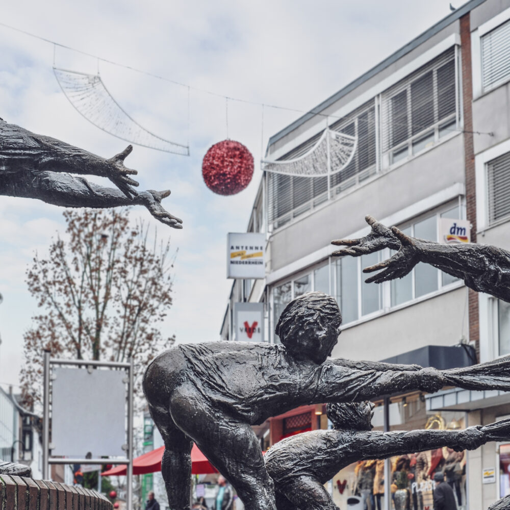 Bronzeskulpturengruppe im Elsabrunnen mit einer Frau, die zwei Kinder an den Händen hält, während ein Engel sie umarmt. Im Hintergrund sind Cafés und Bäume sichtbar.