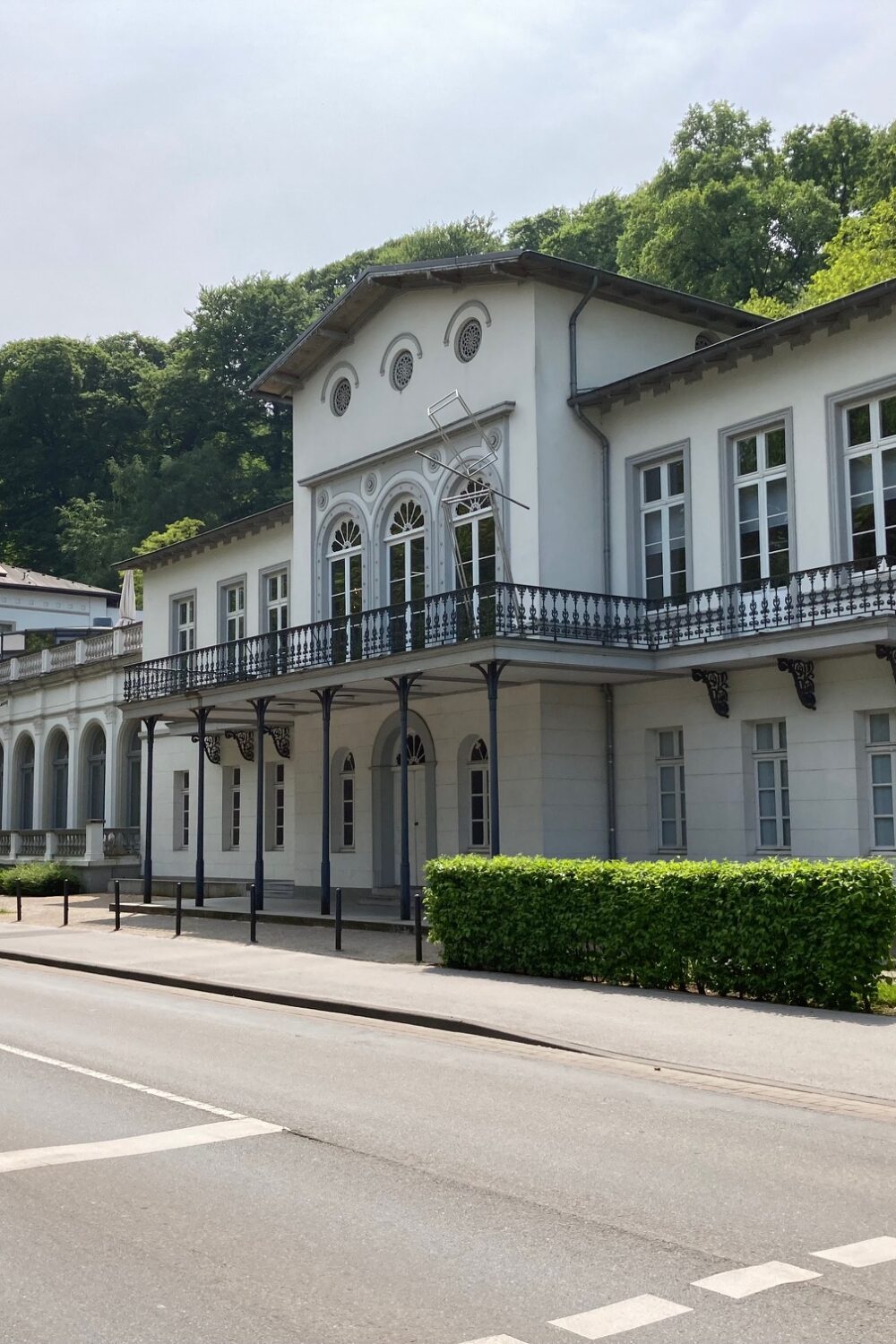 Fassade des Museum Kurhaus Kleve mit weißen Wänden, großen Fenstern und einem Balkon. Im Vordergrund eine Straße mit Bäumen und einem Informationsschild.