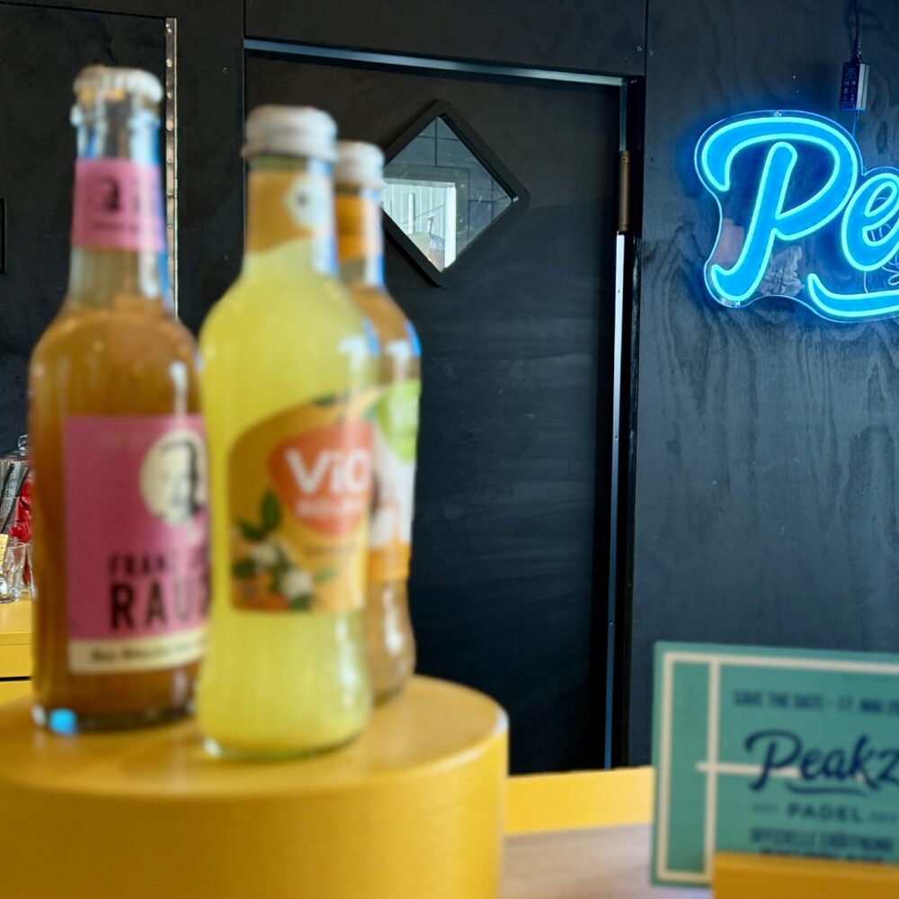 Drei Getränke stehen auf einem gelben Tisch: eine Flasche pinker Limonade, eine Flasche gelber Limonade und eine Flasche mit grünem Etikett. Im Hintergrund leuchtet das Wort 'Peakz' in blauer Neonfarbe.