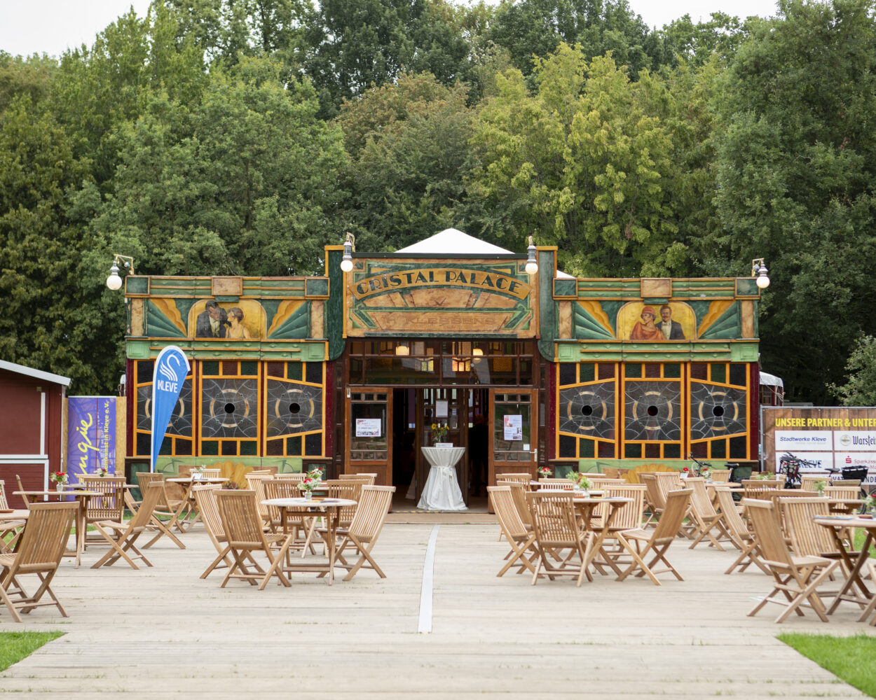Holzterrasse mit vielen leeren Holztischen und -stühlen vor einem grünen, mit Gemälden verzierten Gebäude.