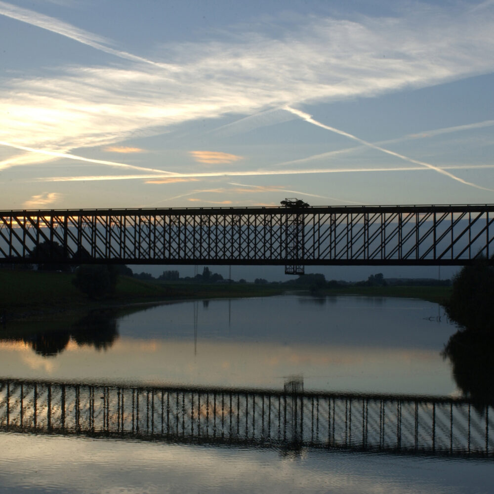 Die alte Bahnbrücke Griethausen ist eine hohe, metallene Fachwerkbrücke, die sich über einen ruhigen Fluss spannt. Ihre Silhouette spiegelt sich im Wasser.