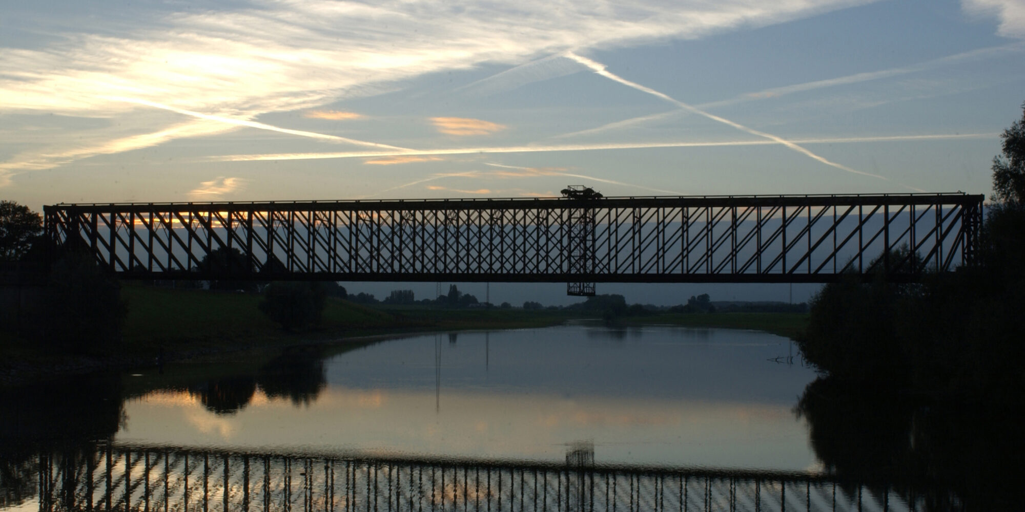 Die alte Bahnbrücke Griethausen ist eine hohe, metallene Fachwerkbrücke, die sich über einen ruhigen Fluss spannt. Ihre Silhouette spiegelt sich im Wasser.