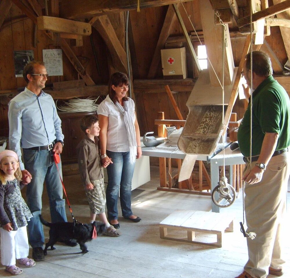Eine Gruppe von vier Personen steht in einer alten Mühle. Ein Mann in grünem Shirt erklärt etwas, während ein Hund an der Leine sitzt. Im Hintergrund sind verschiedene Mühlenteile sichtbar.