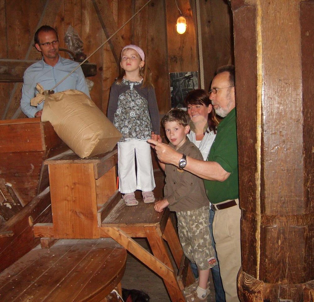 Eine alte Mühle mit Holzstruktur. Ein Kind steht auf einer Holzplattform, während zwei weitere Kinder und ein Erwachsener daneben stehen. Im Hintergrund sind Säcke und Holz sichtbar.