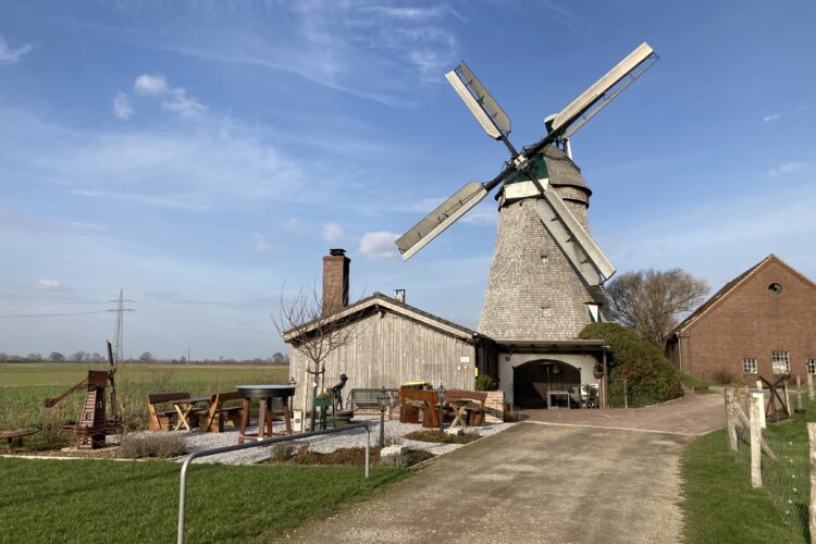 Eine alte Mühle mit grauem Steingebäude und vier großen, beweglichen Flügeln. Im Vordergrund sind Gartenmöbel und eine Statue sichtbar.