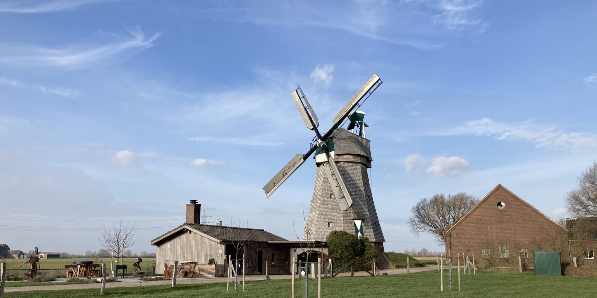 Eine alte Mühle mit grauem Mauerwerk und großen, weißen Flügeln steht in einer grünen Wiese. Neben der Mühle befindet sich ein Nebengebäude aus Holz.