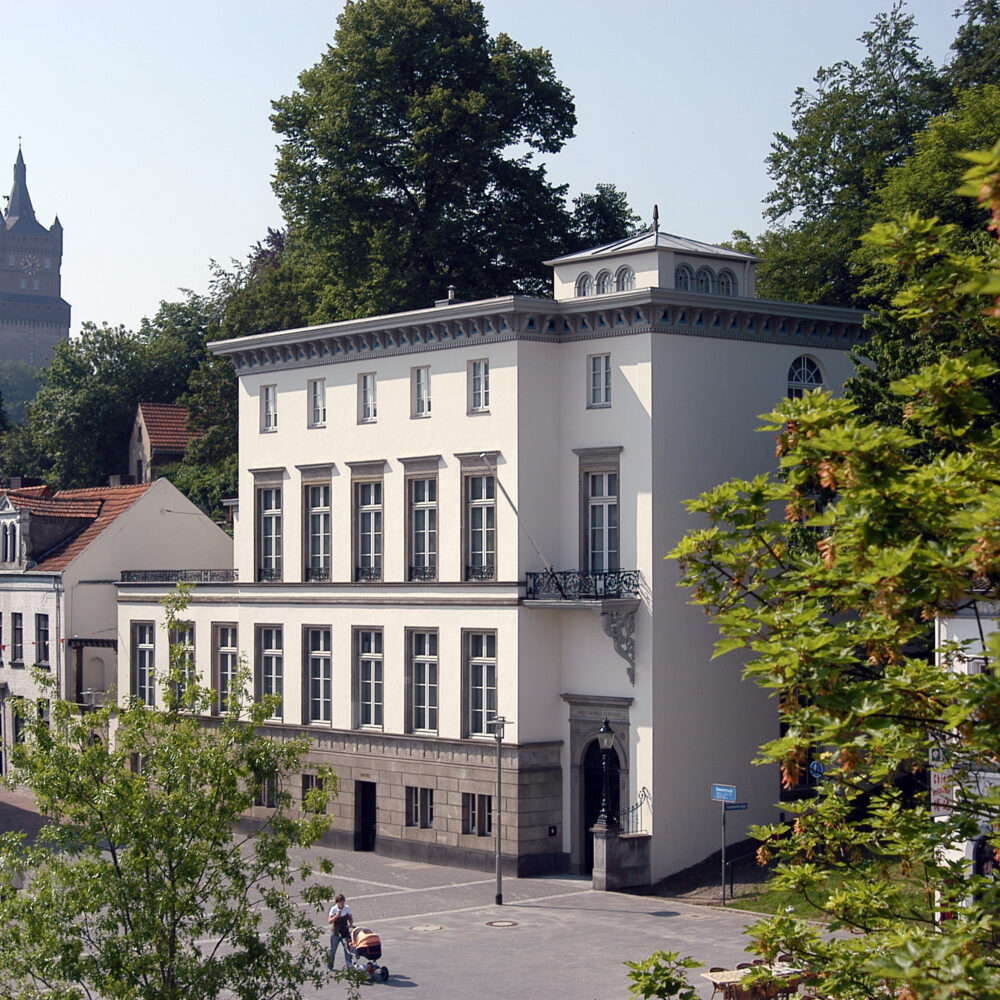 Das B.C. Koekkoek-Haus ist ein dreigeschossiges, weißes Gebäude mit einem Balkon und symmetrischen Fenstern. Im Hintergrund ist die Schwanenburg sichtbar.