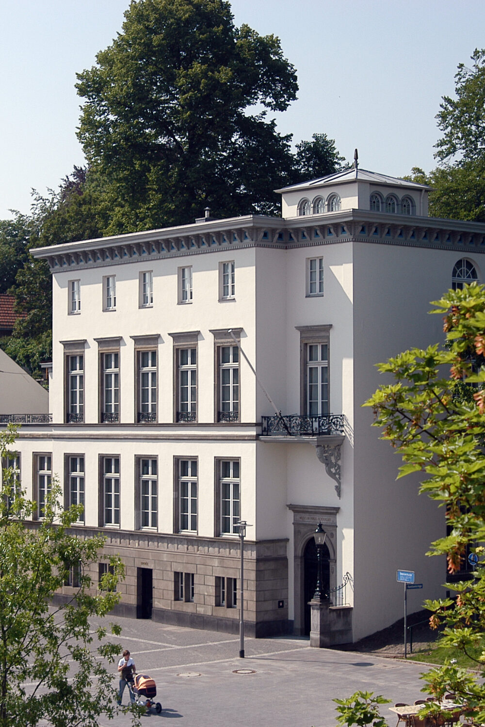 Das B.C. Koekkoek-Haus ist ein dreigeschossiges, weißes Gebäude mit einem Balkon und symmetrischen Fenstern. Im Hintergrund ist die Schwanenburg sichtbar.