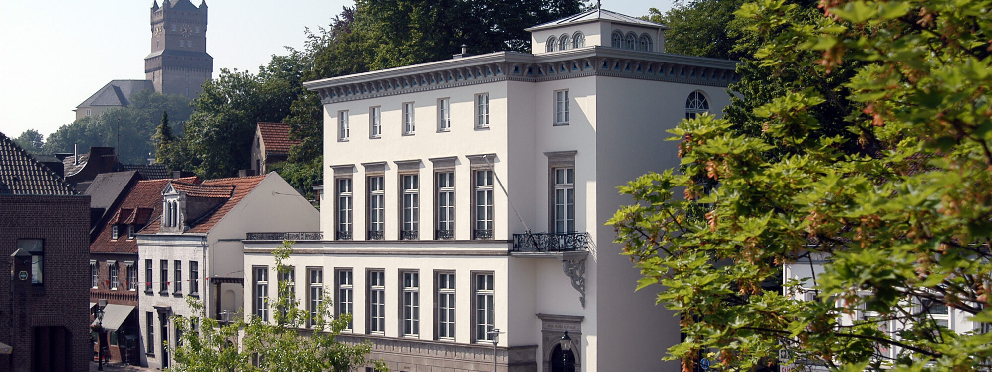 Das B.C. Koekkoek-Haus ist ein dreigeschossiges, weißes Gebäude mit einem Balkon und symmetrischen Fenstern. Im Hintergrund ist die Schwanenburg sichtbar.