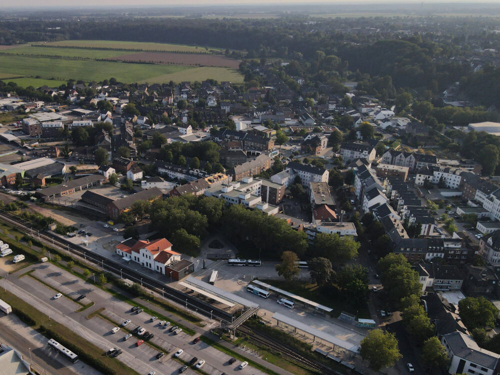 Luftaufnahme von Kleve mit Bahnhof, Parkplätzen, Wohnhäusern und Feldern im Hintergrund bei Tageslicht.