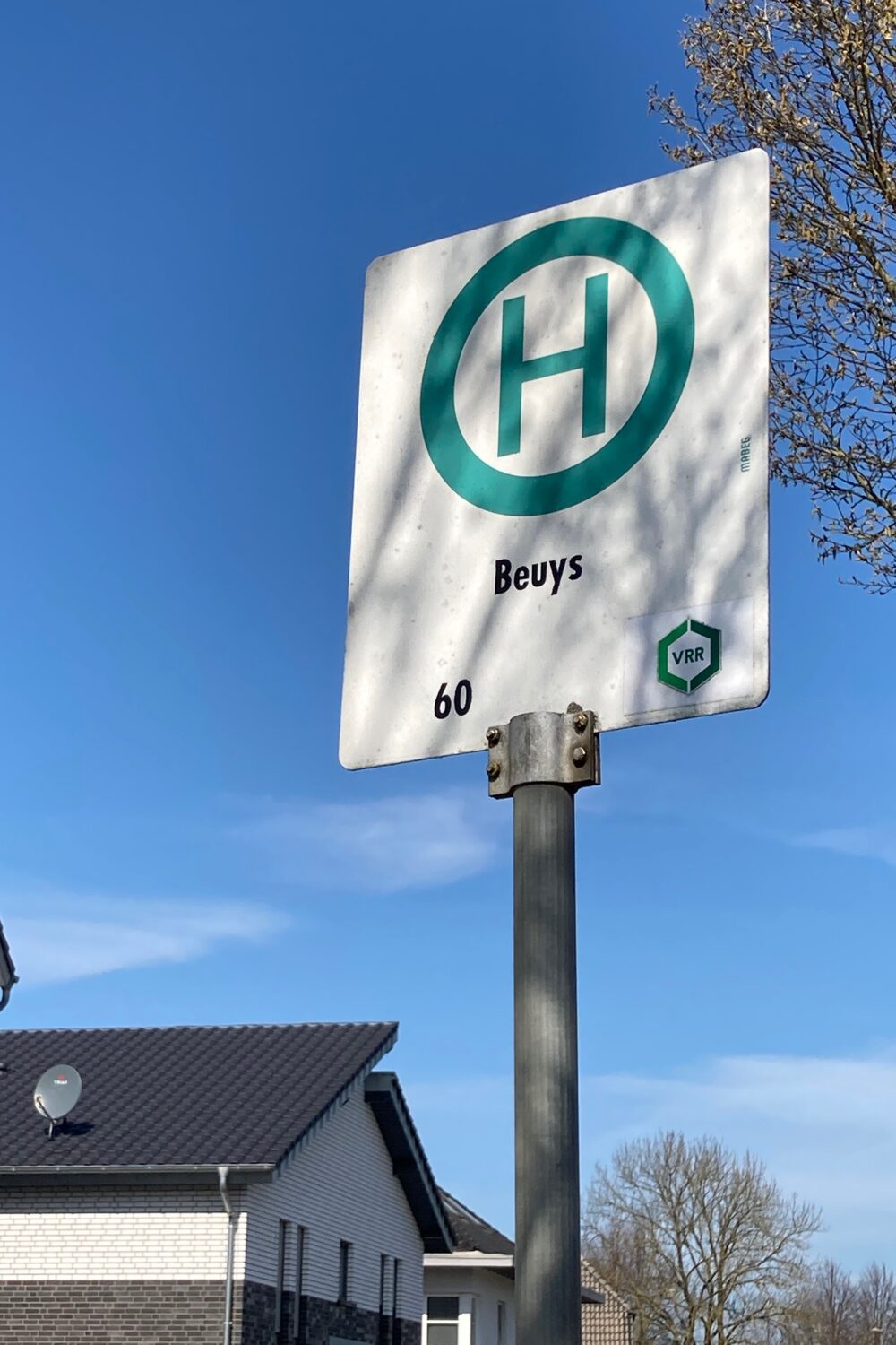 Haltestellenschild mit grünem H und dem Text 'Beuys' sowie der Liniennummer 60. Im Hintergrund ein Wohnhaus und ein Baum.