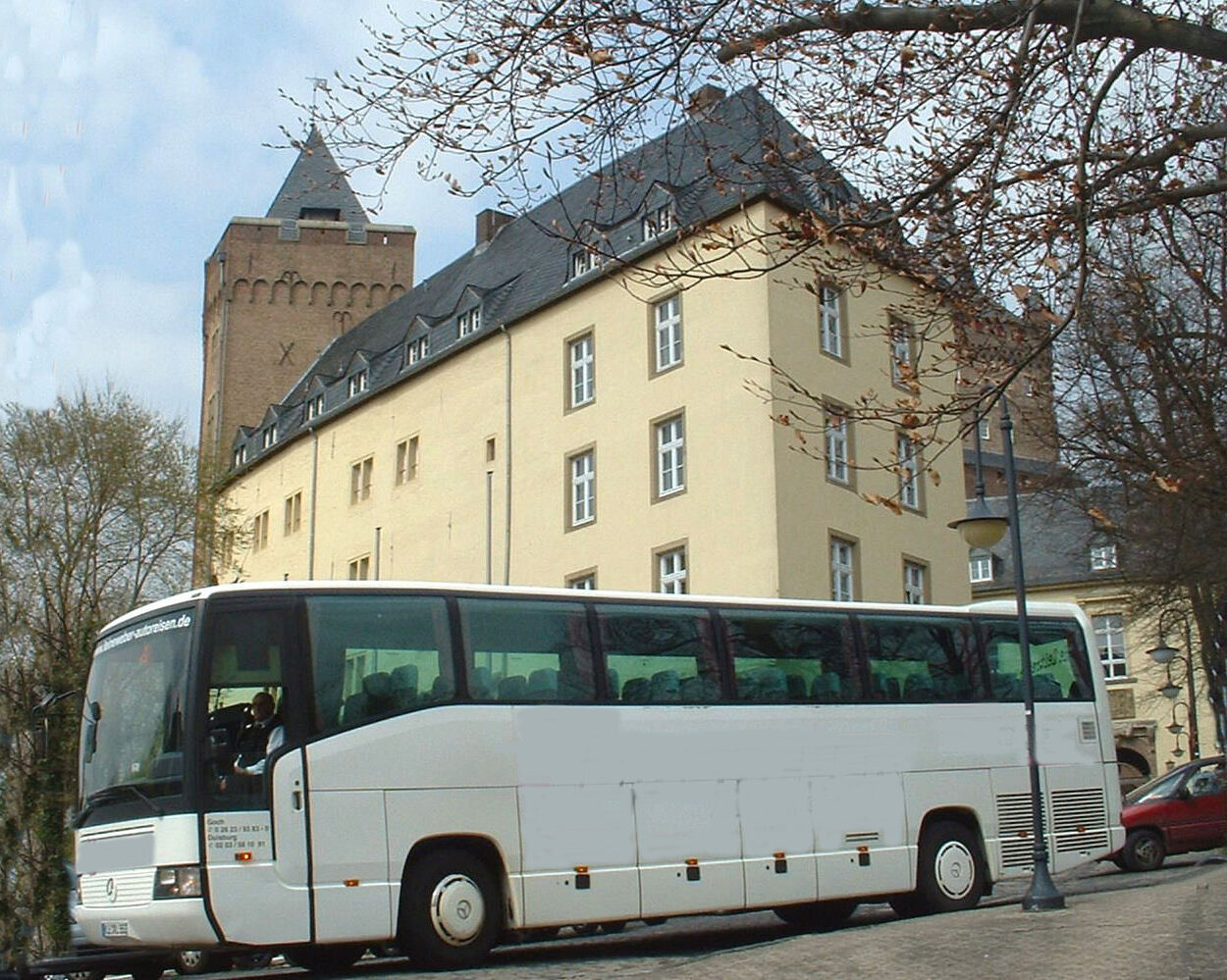 Weißer Reisebus vor der Schwanenburg, im Vordergrund Baumzweige und eine Straßenlaterne