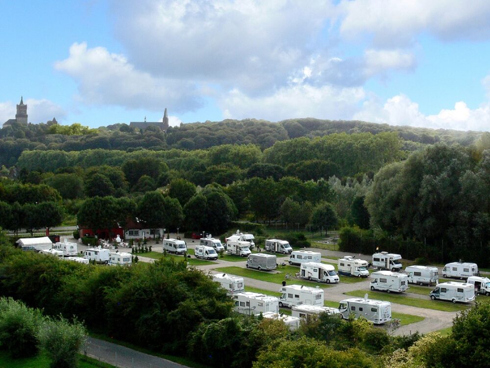 Reisemobil - Campingplatz mit mehreren weißen Wohnmobilen auf grünen Stellplätzen, umgeben von Bäumen und bewaldeten Hügeln mit zwei Kirchtürmen im Hintergrund.