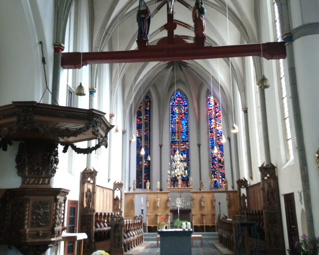 Blick auf den Chorraum der Minoritenkirche mit hohen, spitzbögigen Fenstern und bunten Glasmalereien. In der Mitte steht ein Altar, umgeben von Holzsitzen und einer Kanzel.