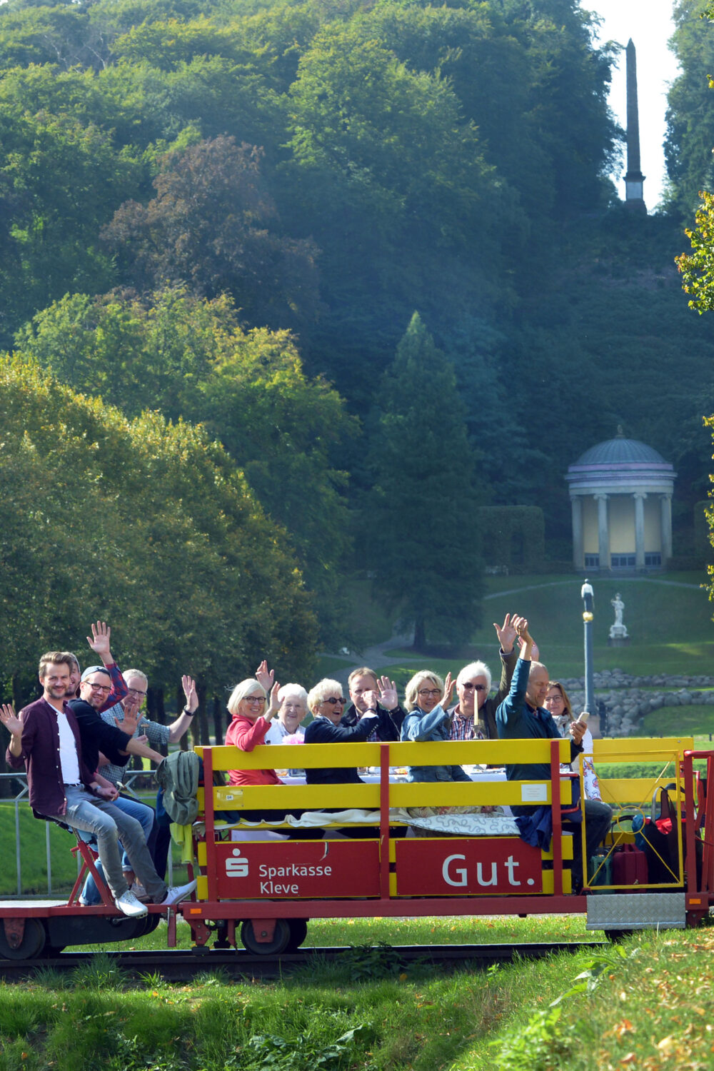 Eine Gruppe von Menschen sitzt auf einer roten Clubdraisine, die auf Schienen fährt. Im Hintergrund sind Bäume und ein Pavillon sichtbar.