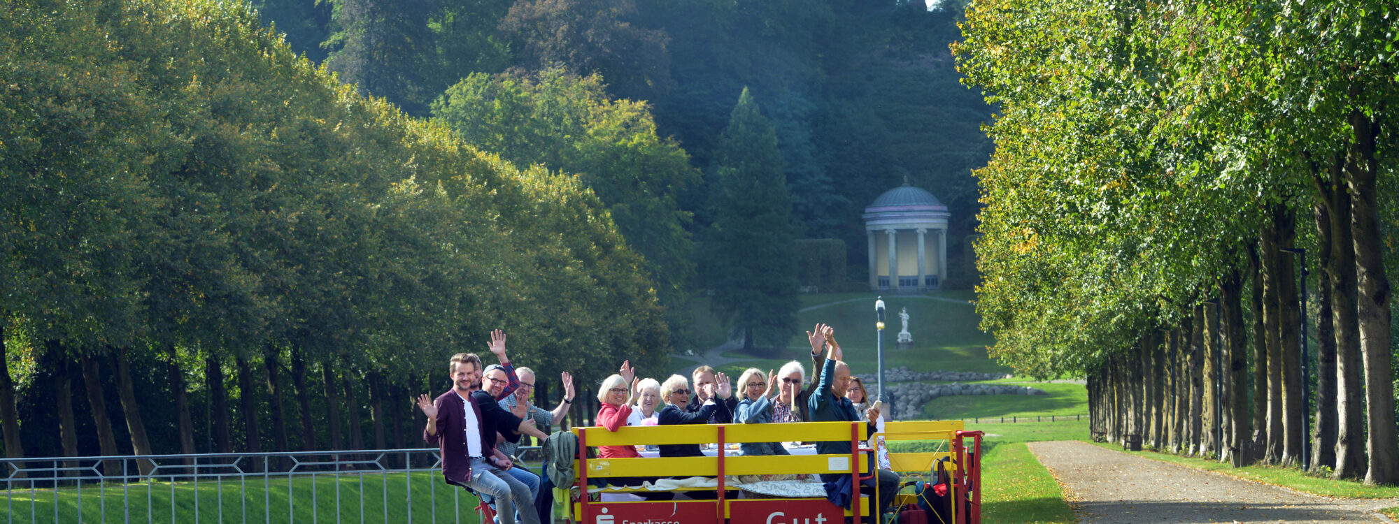 Eine Gruppe von Menschen sitzt auf einer roten Clubdraisine, die auf Schienen fährt. Im Hintergrund sind Bäume und ein Pavillon sichtbar.