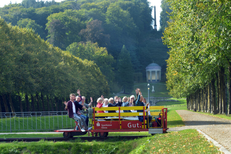 Eine Gruppe von Menschen sitzt auf einer roten Clubdraisine, die auf Schienen fährt. Im Hintergrund sind Bäume und ein Pavillon sichtbar.