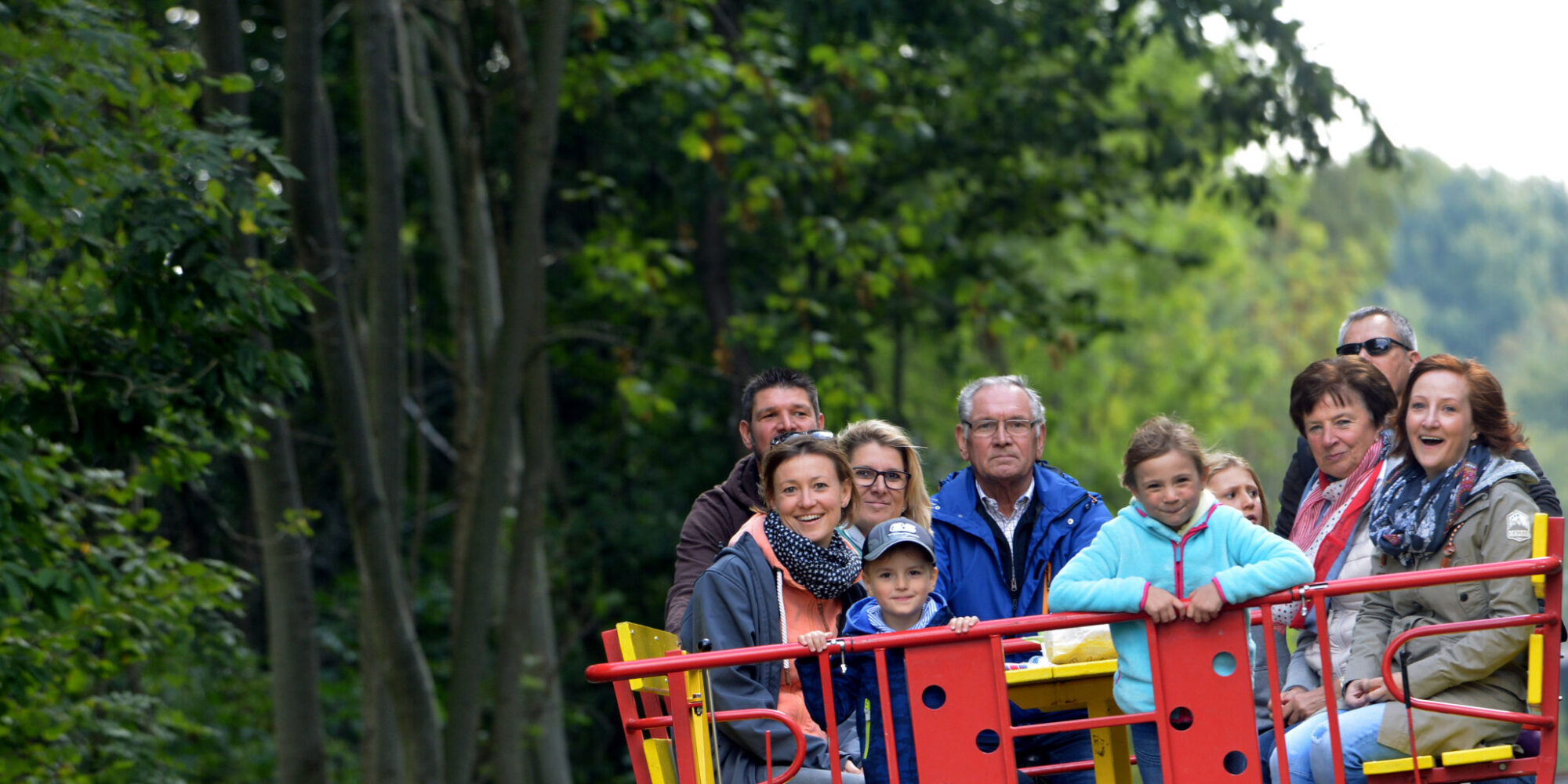 Draisinentour für Familien. Eine Gruppe von zehn Personen sitzt auf einer roten und gelben Clubdraisine, die auf Schienen fährt. Im Hintergrund sind Bäume und ein Stoppschild sichtbar.