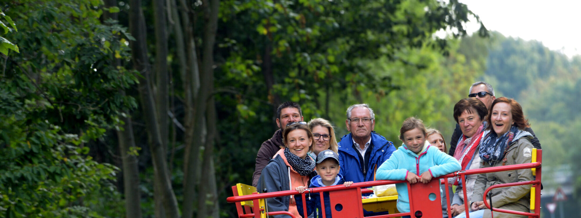 Draisinentour für Familien. Eine Gruppe von zehn Personen sitzt auf einer roten und gelben Clubdraisine, die auf Schienen fährt. Im Hintergrund sind Bäume und ein Stoppschild sichtbar.