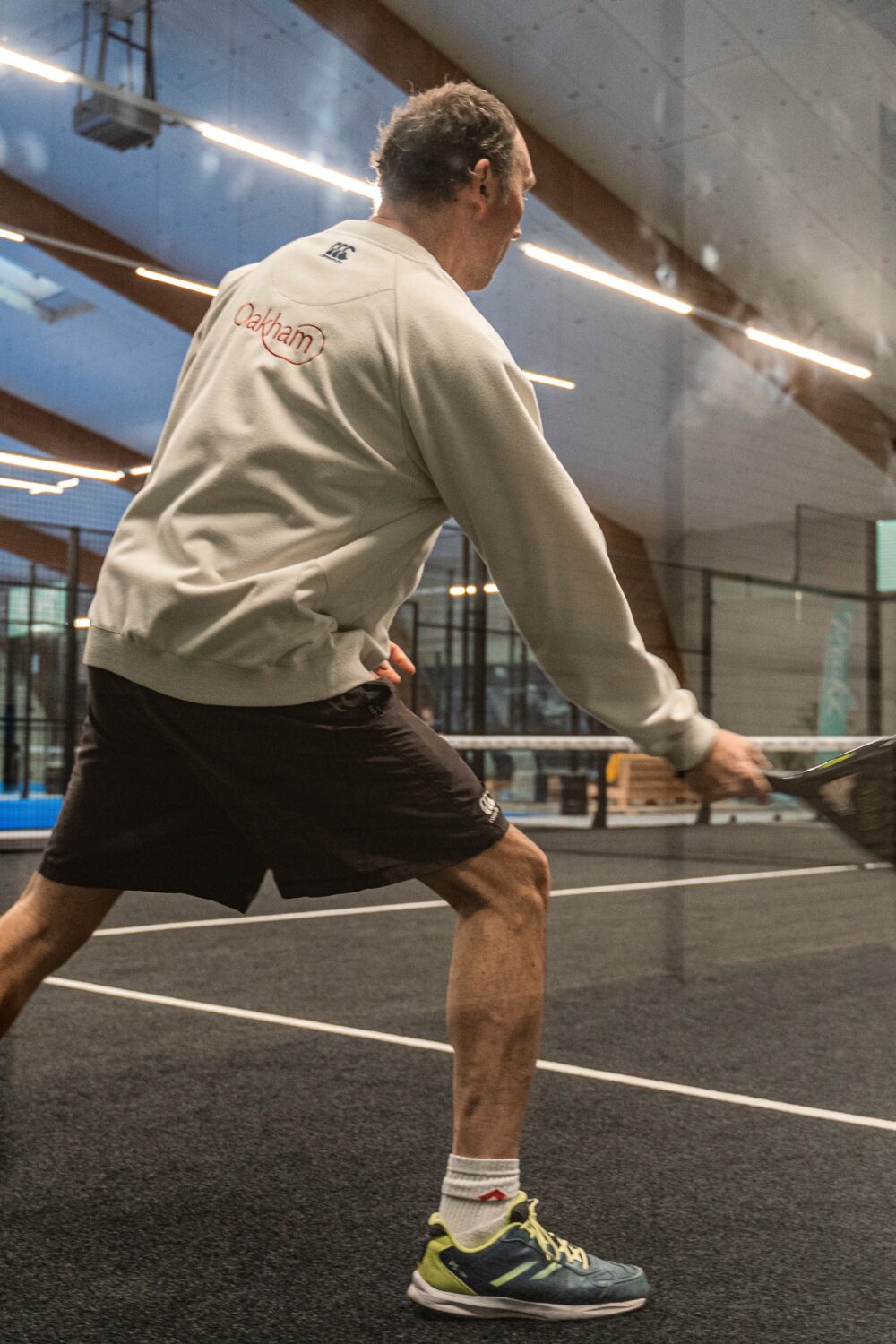 Ein Mann in einem grauen Sweatshirt und schwarzen Shorts schlägt mit einem Schläger einen Tennisball. Im Hintergrund sind weitere Spieler und ein Sportplatz sichtbar.