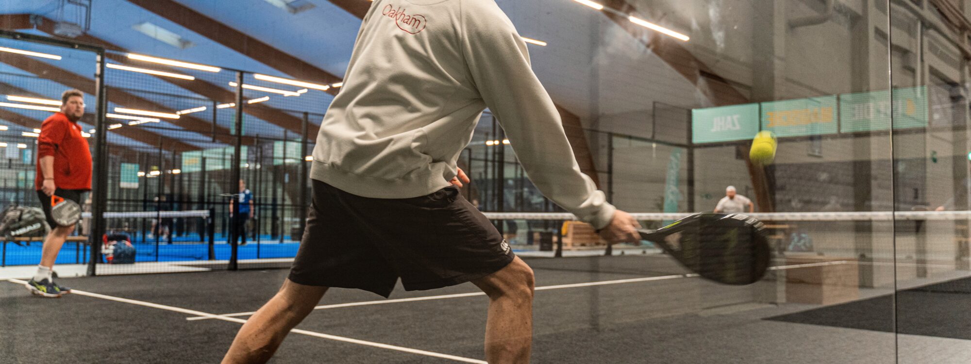 Ein Mann in einem grauen Sweatshirt und schwarzen Shorts schlägt mit einem Schläger einen Tennisball. Im Hintergrund sind weitere Spieler und ein Sportplatz sichtbar.