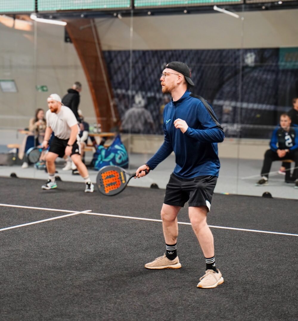 Zwei Personen spielen Padel-Tennis in einer Halle. Eine Person in blauer Jacke und schwarzer Shorts steht bereit, während die andere Person im Hintergrund einen Schlag ausführt.