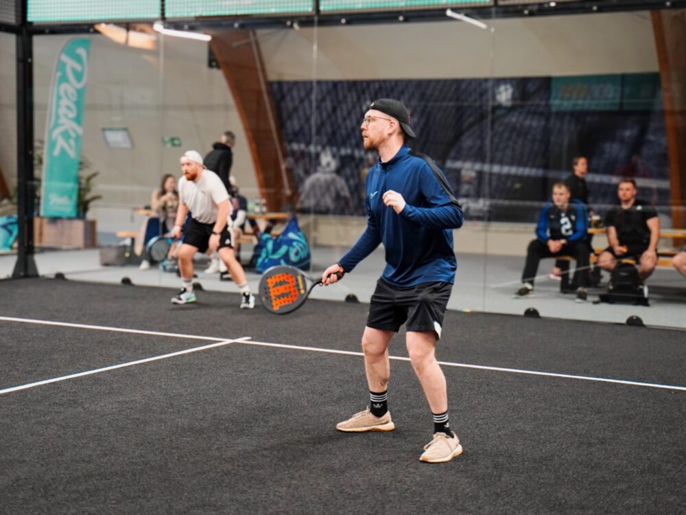 Zwei Personen spielen Padel-Tennis in einer Halle. Eine Person in blauer Jacke und schwarzer Shorts steht bereit, während die andere Person im Hintergrund einen Schlag ausführt.