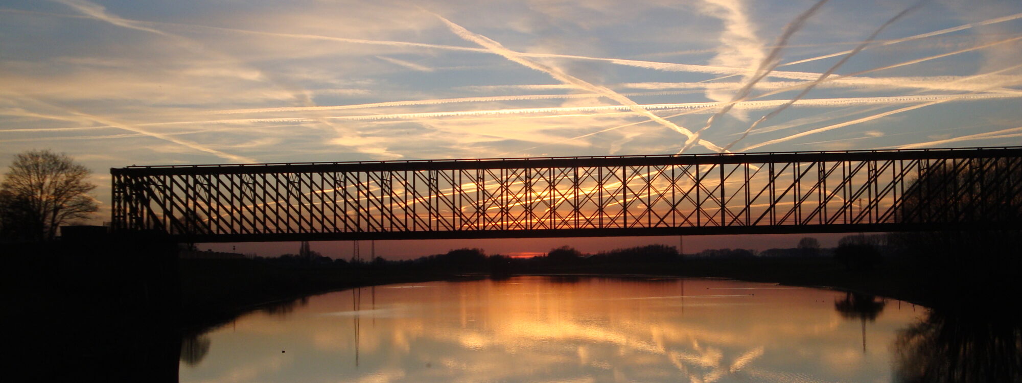 Die alte Bahnbrücke Griethausen ist eine hohe, metallene Fachwerkbrücke, die sich über einen ruhigen Fluss spannt. Ihre Silhouette spiegelt sich im Wasser.