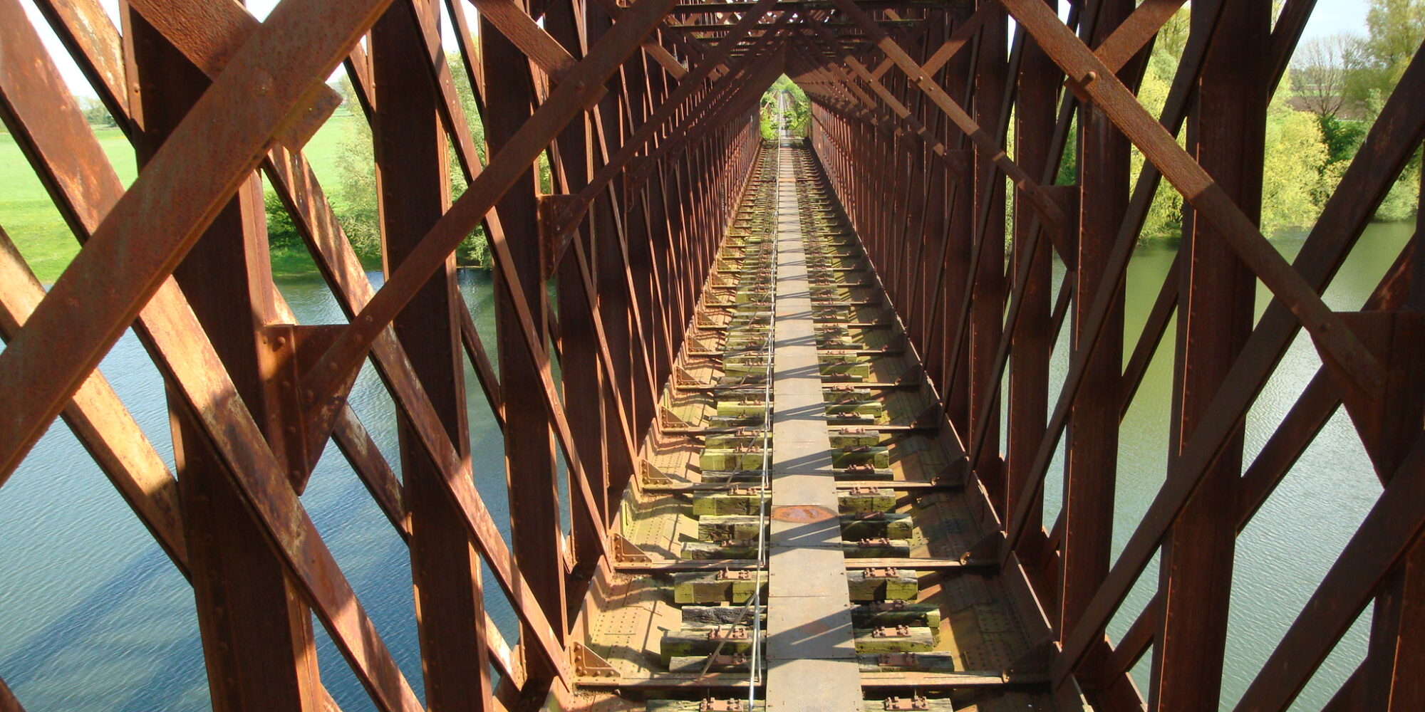 Blick auf eine alte, rostige Eisenbahnbrücke mit sichtbaren Stahlträgern und einem schmalen Weg in der Mitte, der über Wasser führt.