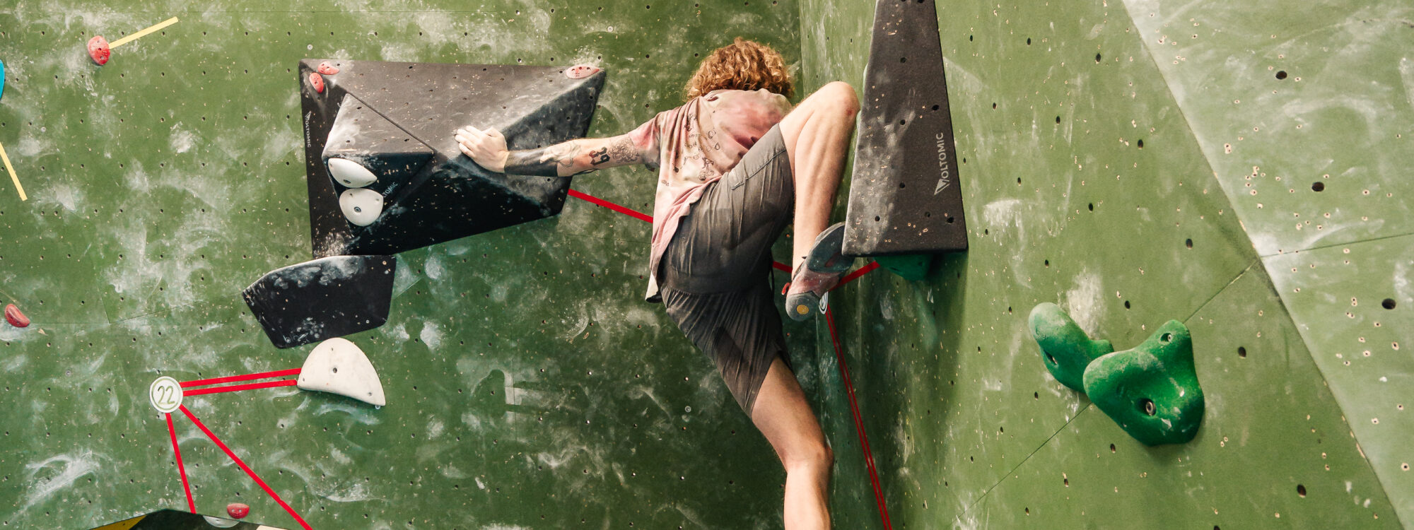 Ein Kletterer in kurzer Hose und T-Shirt klettert an einer grünen Kletterwand mit schwarzen und grünen Griffen. Im Hintergrund sind weitere Kletterer zu sehen.