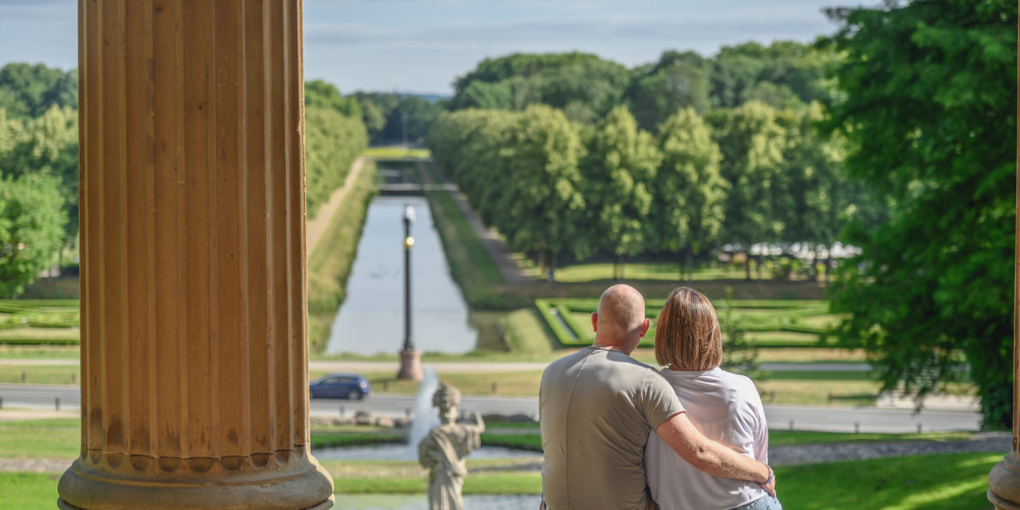 Pärchen sitzt auf einer Treppe und schaut auf die Gartenanlagen mit dem Neuer Eiserner Mann, einem Wasserlauf und Bäumen im Hintergrund. Der Mann hat eine Glatze und trägt ein graues T-Shirt, die Frau hat kurze Haare und trägt eine weiße Bluse.