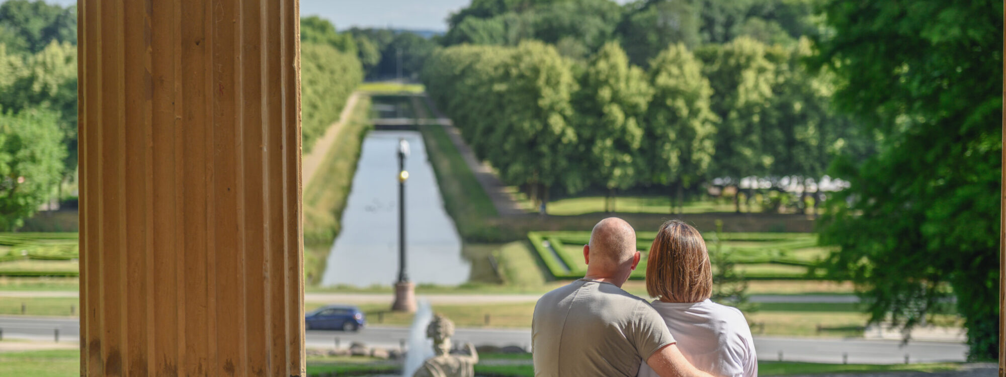 Pärchen sitzt auf einer Treppe und schaut auf die Gartenanlagen mit dem Neuer Eiserner Mann, einem Wasserlauf und Bäumen im Hintergrund. Der Mann hat eine Glatze und trägt ein graues T-Shirt, die Frau hat kurze Haare und trägt eine weiße Bluse.