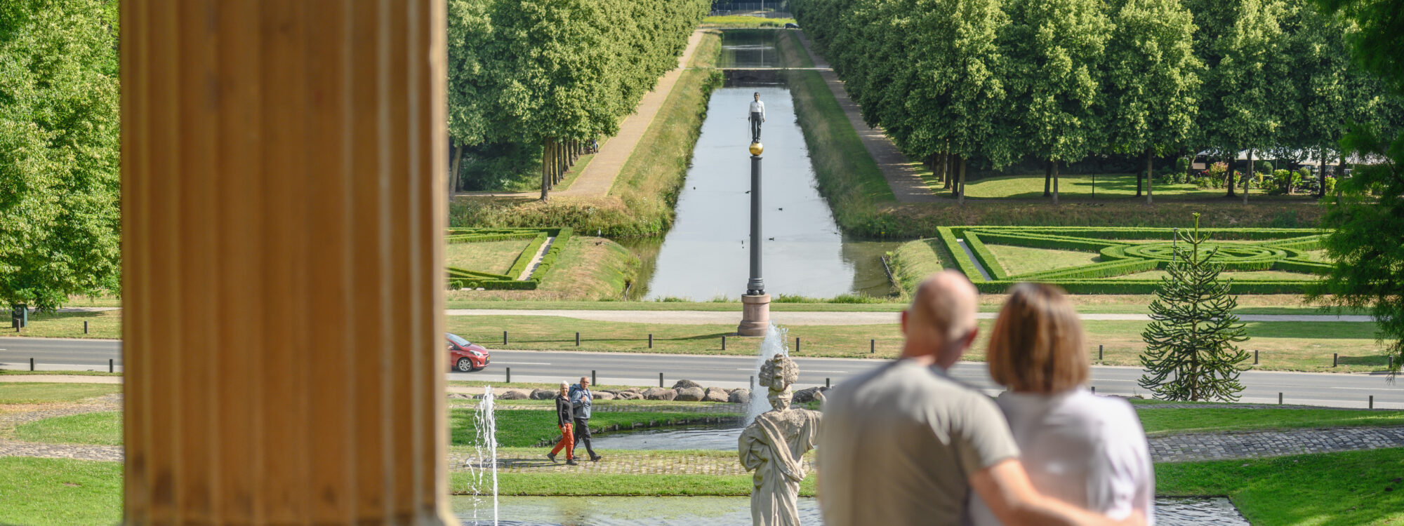 Pärchen sitzt auf einer Treppe und schaut auf die Gartenanlagen mit einem Wasserlauf und Bäumen im Hintergrund. Der Mann hat eine Glatze und trägt ein graues T-Shirt, die Frau hat kurze Haare und trägt eine weiße Bluse.