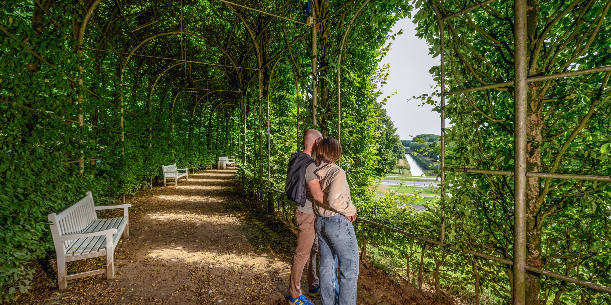 Paar steht umarmt in einem begrünten Laubengang in den Gartenanlagen. Im Hintergrund sind zwei weiße Bänke und eine Landschaft sichtbar.