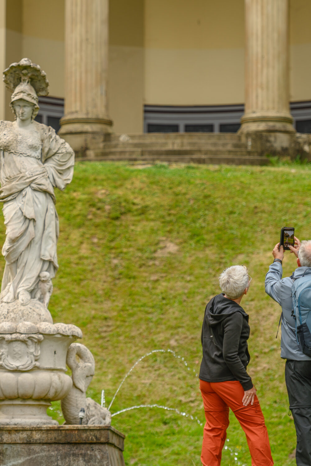 Eine Statue einer weiblichen Figur mit einer Schale auf dem Kopf steht auf einem Brunnen. Im Hintergrund sind drei Personen zu sehen, die ein Foto in den Gartenanlagen machen.