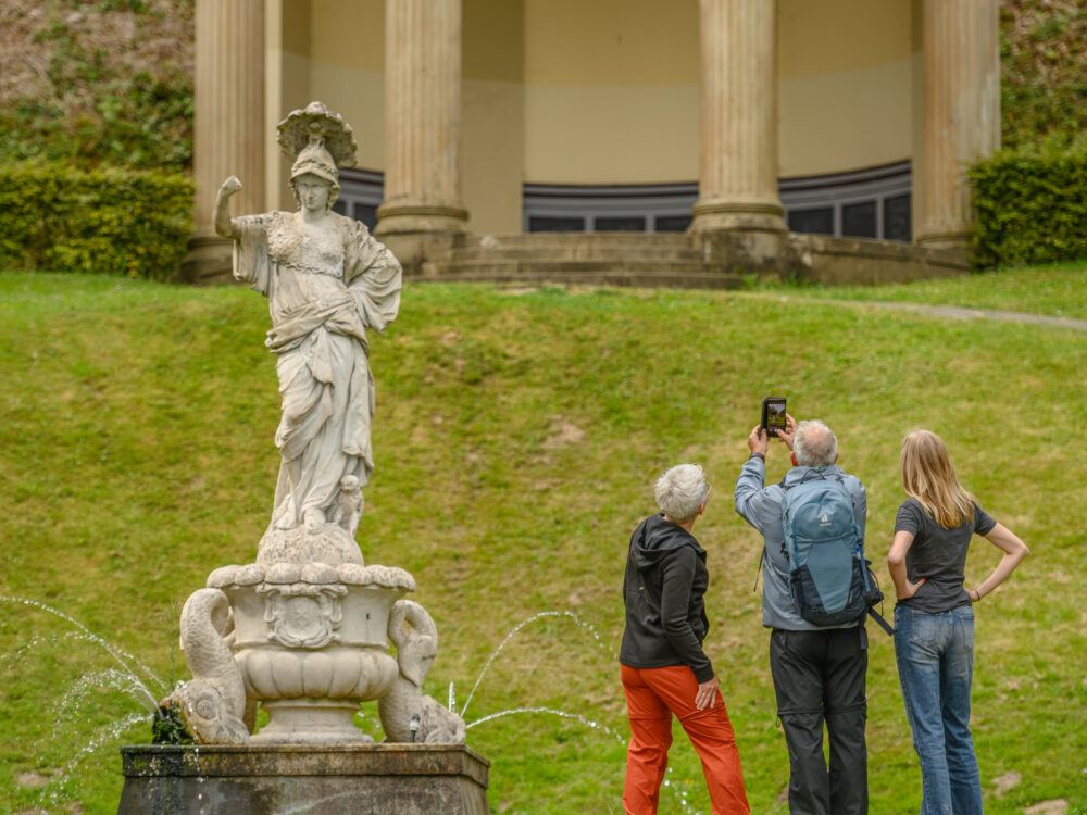 Eine Statue einer weiblichen Figur mit einer Schale auf dem Kopf steht auf einem Brunnen. Im Hintergrund sind drei Personen zu sehen, die ein Foto in den Gartenanlagen machen.