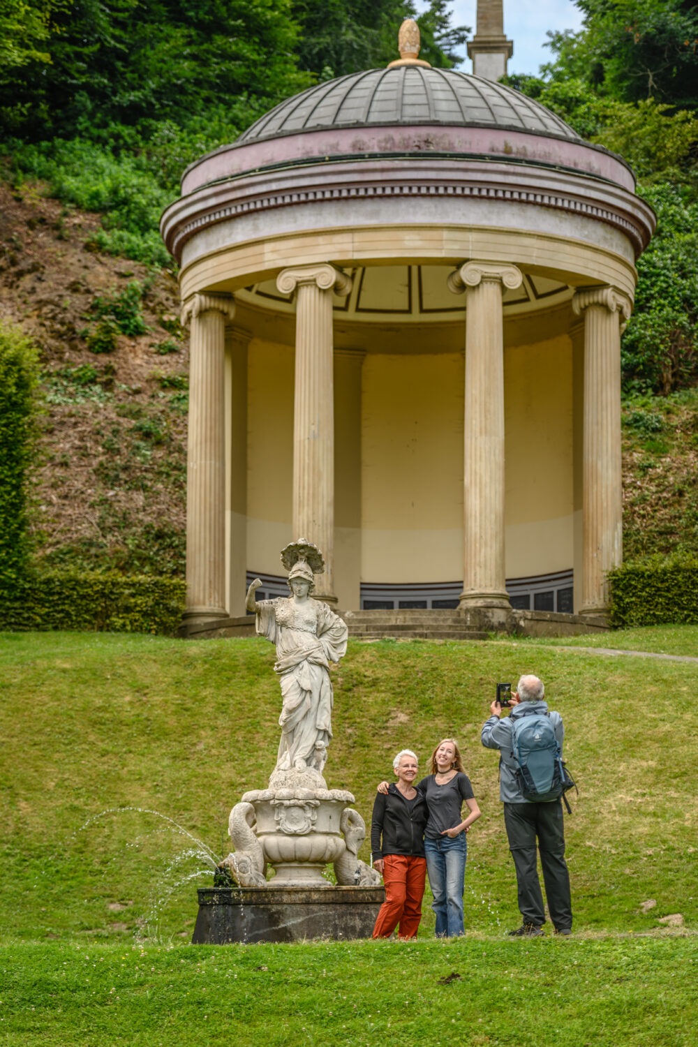 Runder Pavillon in den Gartenanlagen mit Kuppeldach und vier Säulen auf einem Hügel, davor eine steinerne Statue mit Wasserspielen und drei Personen, von denen eine fotografiert.