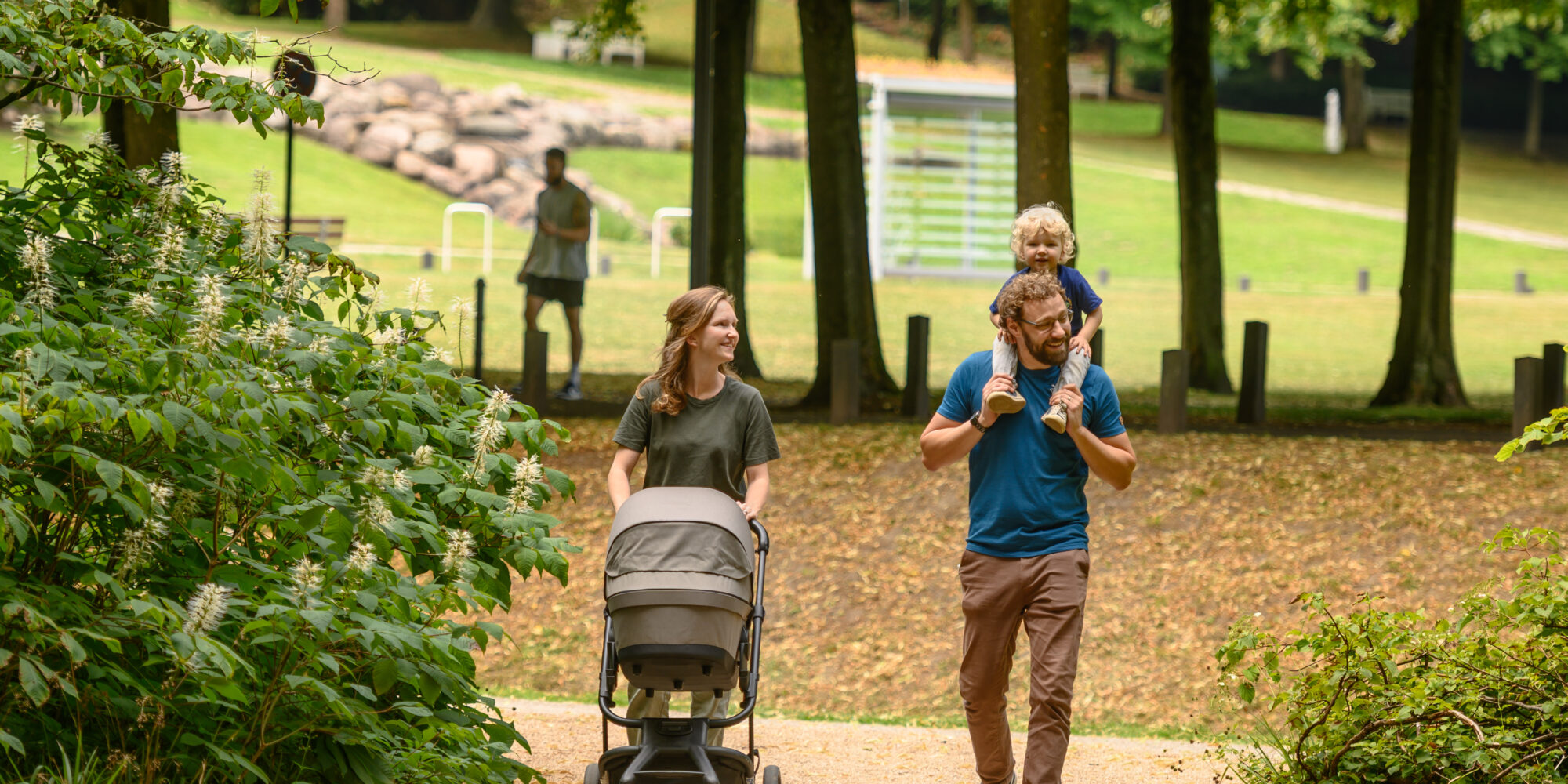 Eine Frau schiebt einen Kinderwagen auf einem Weg, während ein Mann ein Kind auf den Schultern trägt. Im Hintergrund ist eine Person zu sehen, die in einem Park steht.