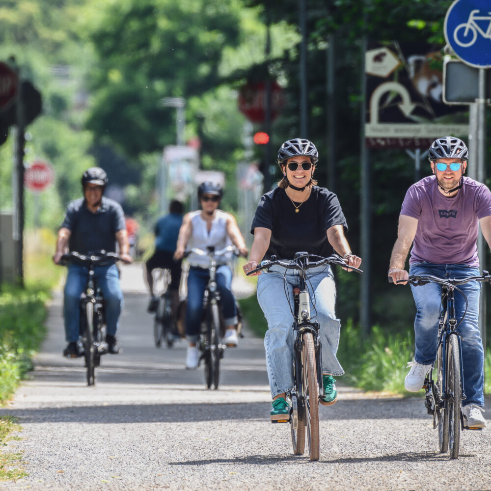 Radfahren in Kleve. Fünf Personen auf Fahrrädern fahren auf einem Radweg. Vier Personen tragen Helme, eine Person fährt ein E-Bike. Verkehrsschilder sind im Hintergrund sichtbar.