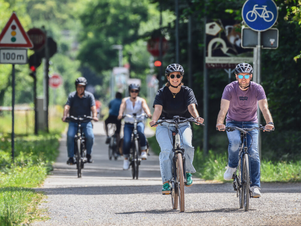 Radfahren in Kleve. Fünf Personen auf Fahrrädern fahren auf einem Radweg. Vier Personen tragen Helme, eine Person fährt ein E-Bike. Verkehrsschilder sind im Hintergrund sichtbar.