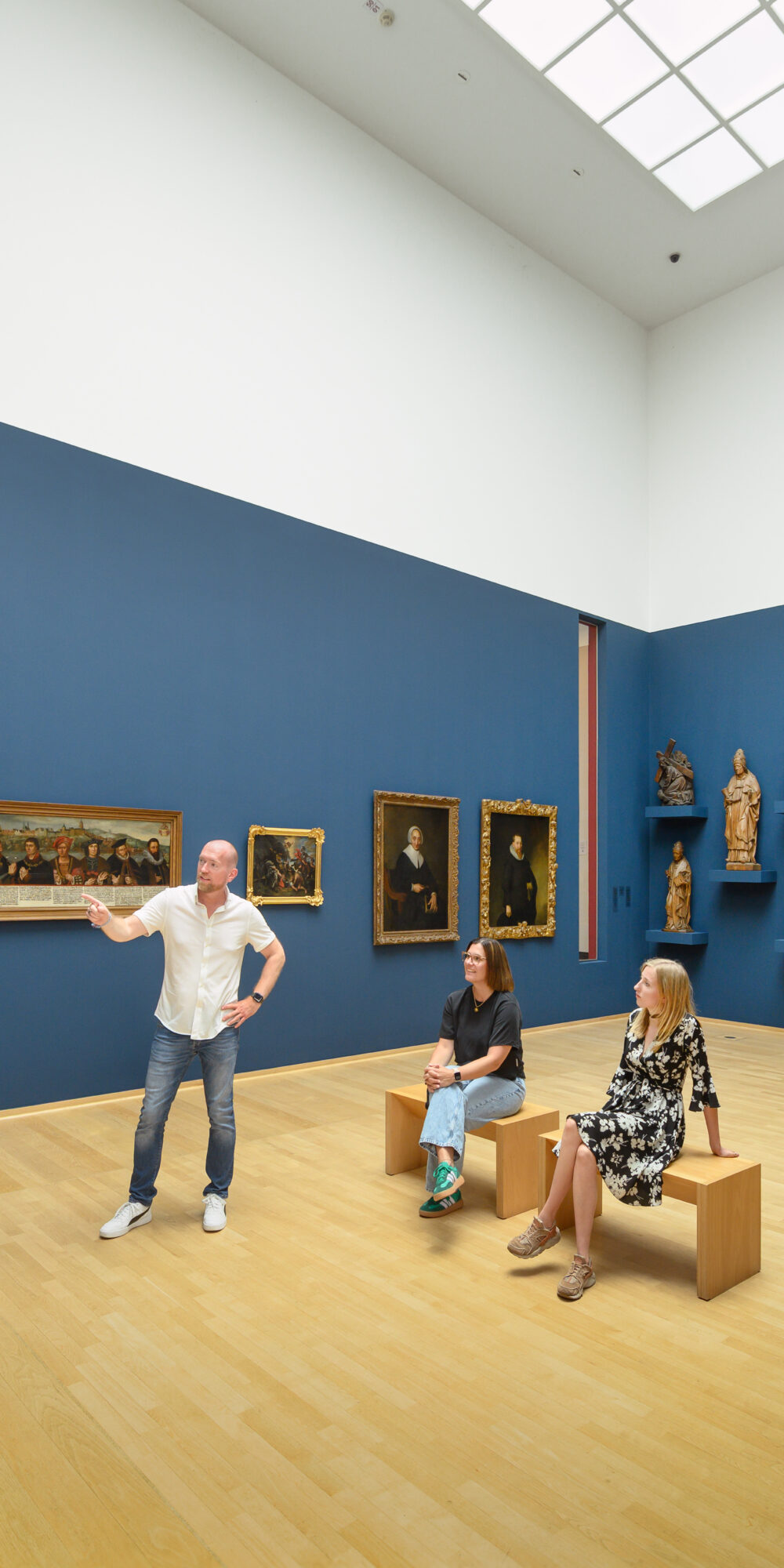 Drei Personen sitzen und stehen im Museum Kurhaus Kleve in einem Museumssaal mit blauen Wänden, auf denen Gemälde und Holzskulpturen ausgestellt sind.