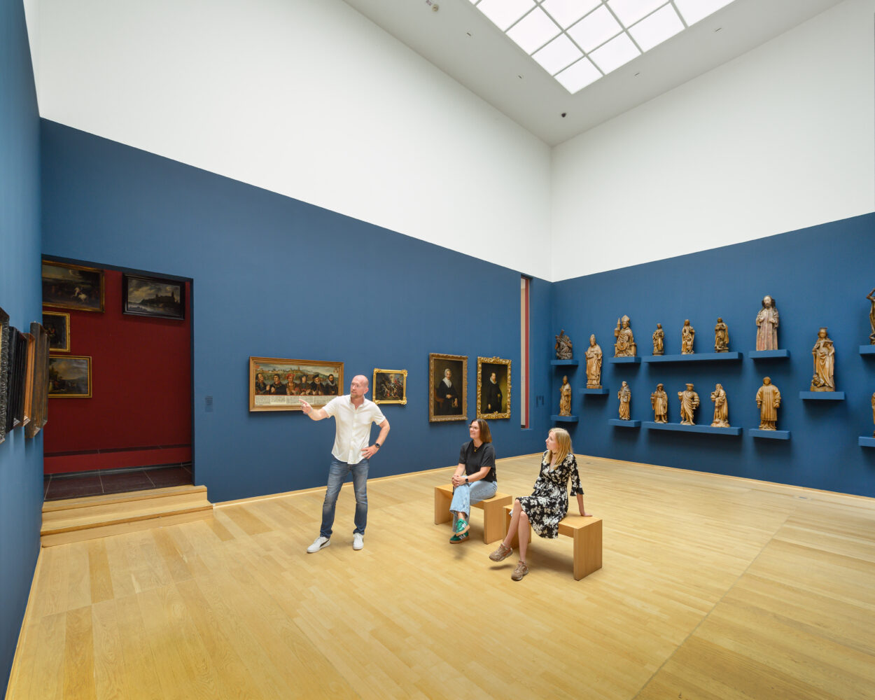 Drei Personen sitzen und stehen im Museum Kurhaus Kleve in einem Museumssaal mit blauen Wänden, auf denen Gemälde und Holzskulpturen ausgestellt sind.