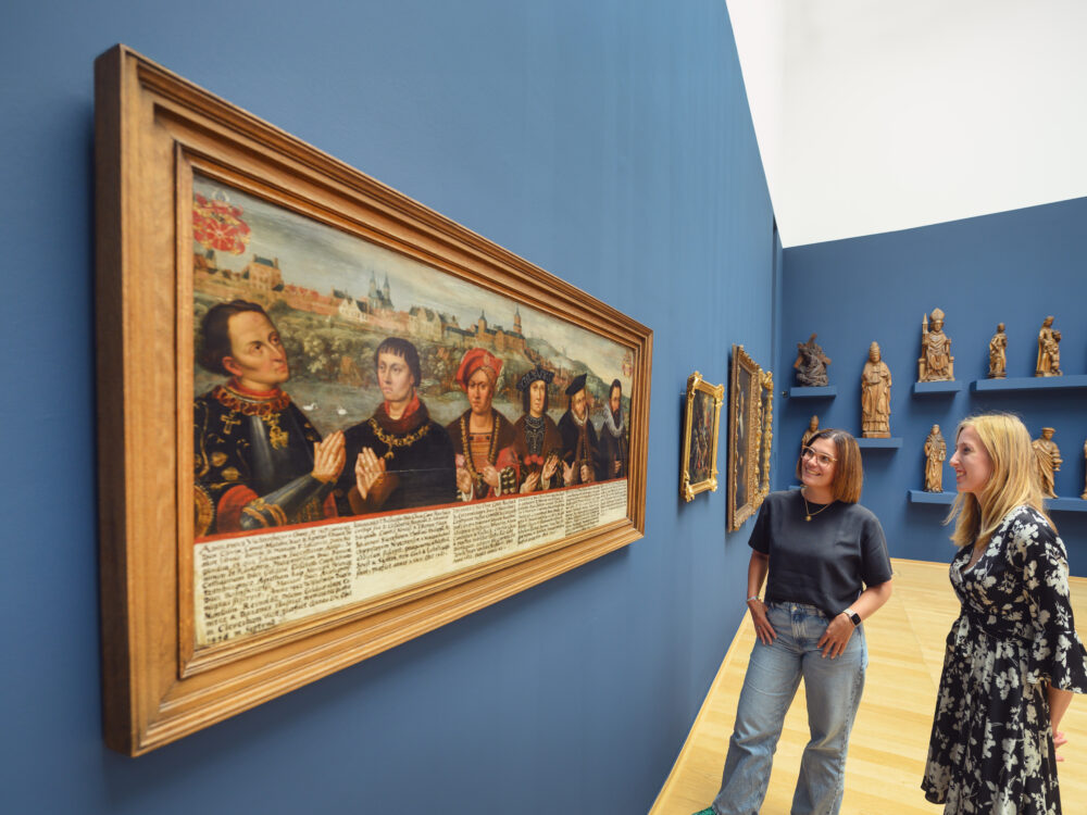Zwei Personen stehen vor einem großen Gemälde mit historischen Figuren und einer beschrifteten Unterzeile. Im Hintergrund sind Holzskulpturen an der Wand zu sehen.