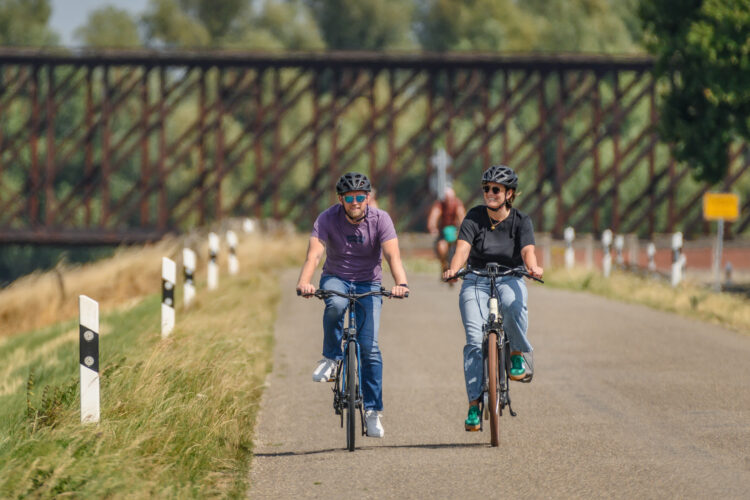 Zwei Radfahrer auf einer Straße, einer in lila T-Shirt und einer in schwarzem T-Shirt, beide mit Helmen. Im Hintergrund eine Eisenbahnbrücke.