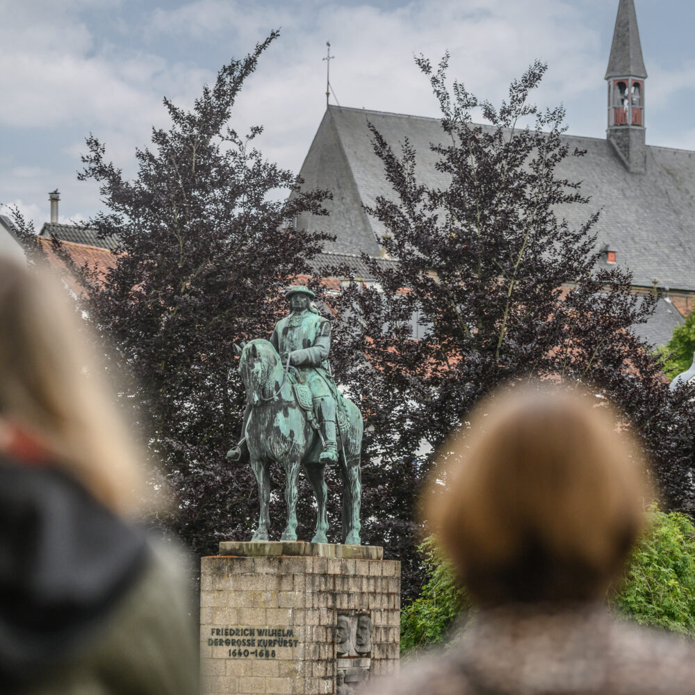 Der Große Kurfürst Friedrich Wilhelm als Bronzestatue auf einem Steinsockel. Zwei Frauen blicken auf das Denkmal des Großen Kurfürsten. Bäume und andere Gebäude sind im Hintergrund.
