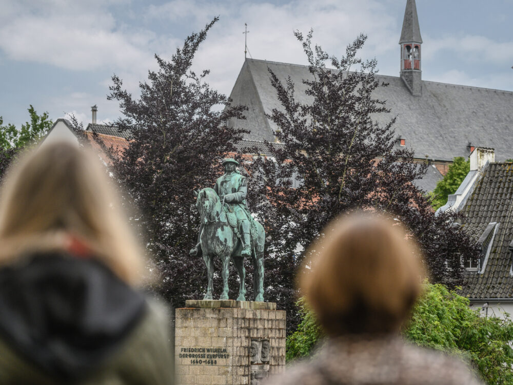 Der Große Kurfürst Friedrich Wilhelm als Bronzestatue auf einem Steinsockel. Zwei Frauen blicken auf das Denkmal des Großen Kurfürsten. Bäume und andere Gebäude sind im Hintergrund.