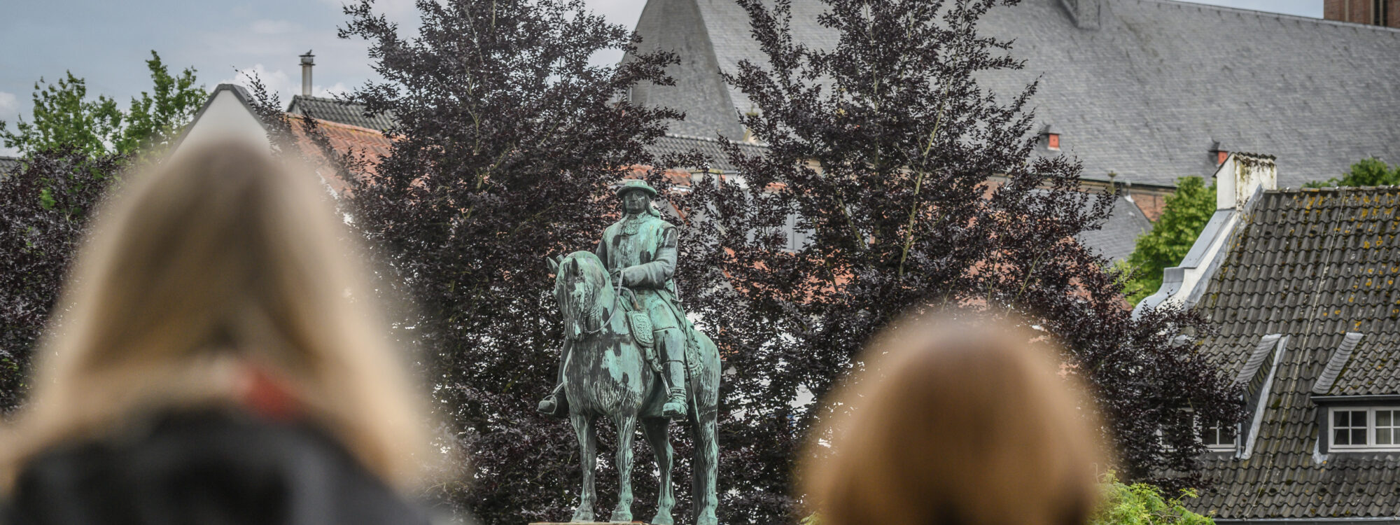 Der Große Kurfürst Friedrich Wilhelm als Bronzestatue auf einem Steinsockel. Zwei Frauen blicken auf das Denkmal des Großen Kurfürsten. Bäume und andere Gebäude sind im Hintergrund.