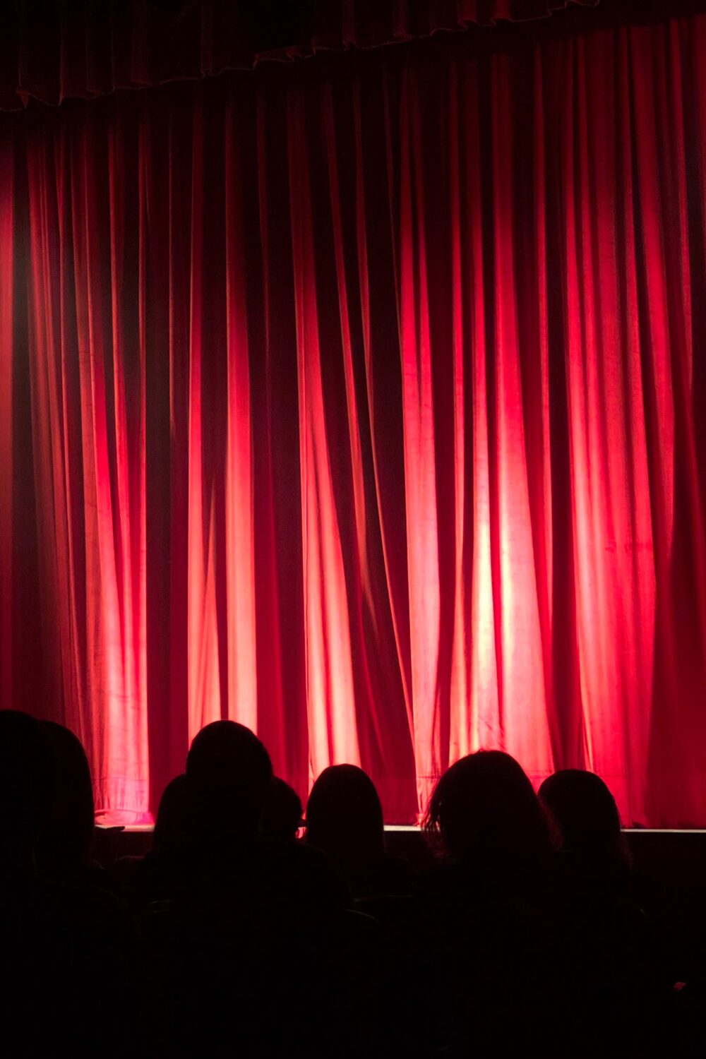 Rote Theaterbühne mit geschlossenen Vorhängen und Silhouetten von Zuschauern im Vordergrund.
