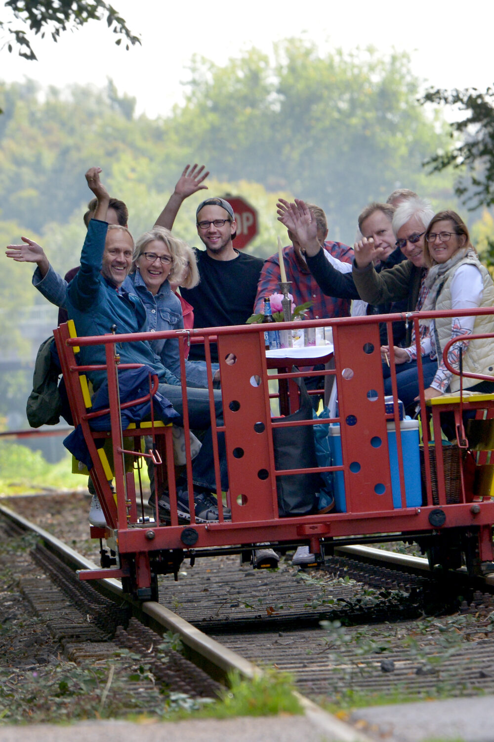 Eine rote Draisine mit mehreren Personen, die auf den Sitzen sitzen und die Hände heben. Im Hintergrund sind Bäume und ein Verkehrsschild mit der Aufschrift '100 m' sichtbar.